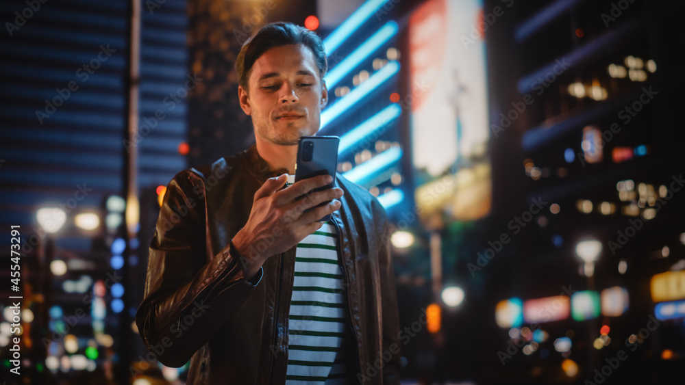 Man Using Smartphone Walking Through Night City Street Full of Neon Light. Smiling Stylish Man Using