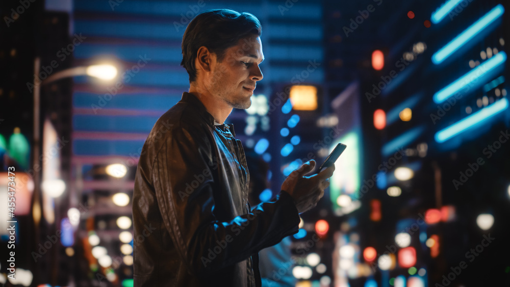 手持智能手机的英俊年轻人站在霓虹灯下的夜城街道上的肖像