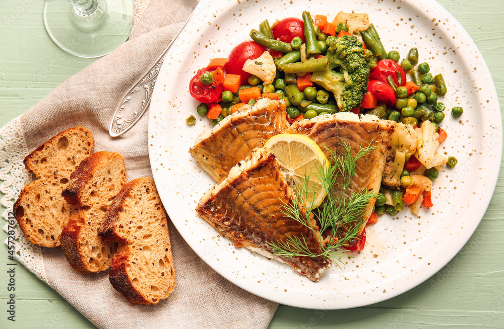 彩色木质背景上有美味的烤鳕鱼片、蔬菜和面包