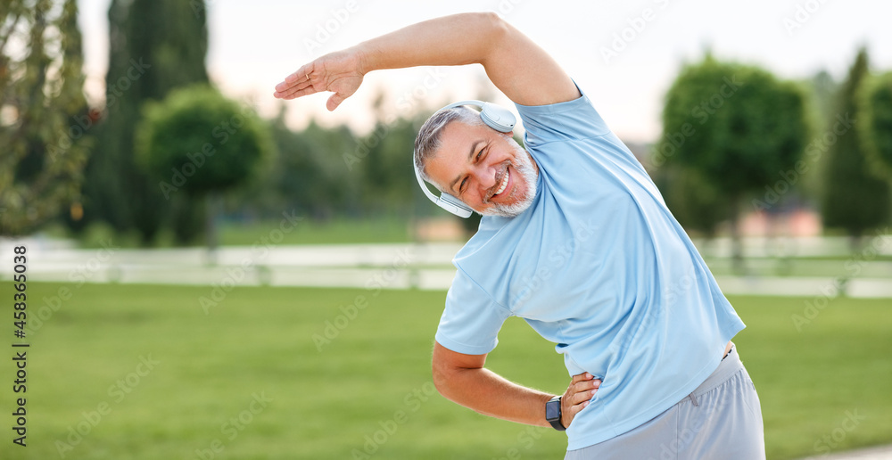Happy retired senior man doing side stretching exercises with arm overhead during outdoor workout