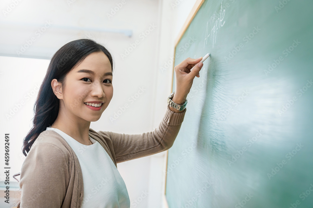 年轻快乐的亚洲老师在黑板上写字的特写肖像。女人在黑板墙上写字