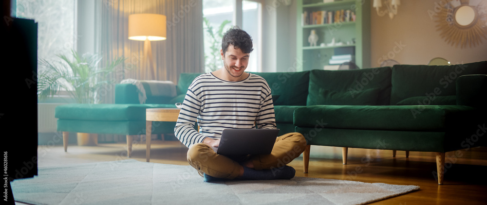 英俊的成年男子坐在舒适时尚的公寓客厅地板上使用笔记本电脑