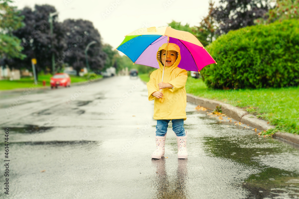 穿着黄色防水外套和靴子，拿着五颜六色的雨伞，在t中玩耍的滑稽可爱的小女孩
