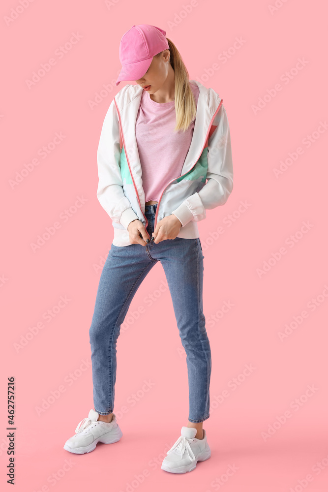 戴棒球帽的时尚女性在粉色背景下拉拉链运动衫