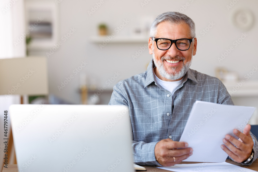 .一位英俊微笑的老人在家里用笔记本电脑远程工作，阅读财务文件