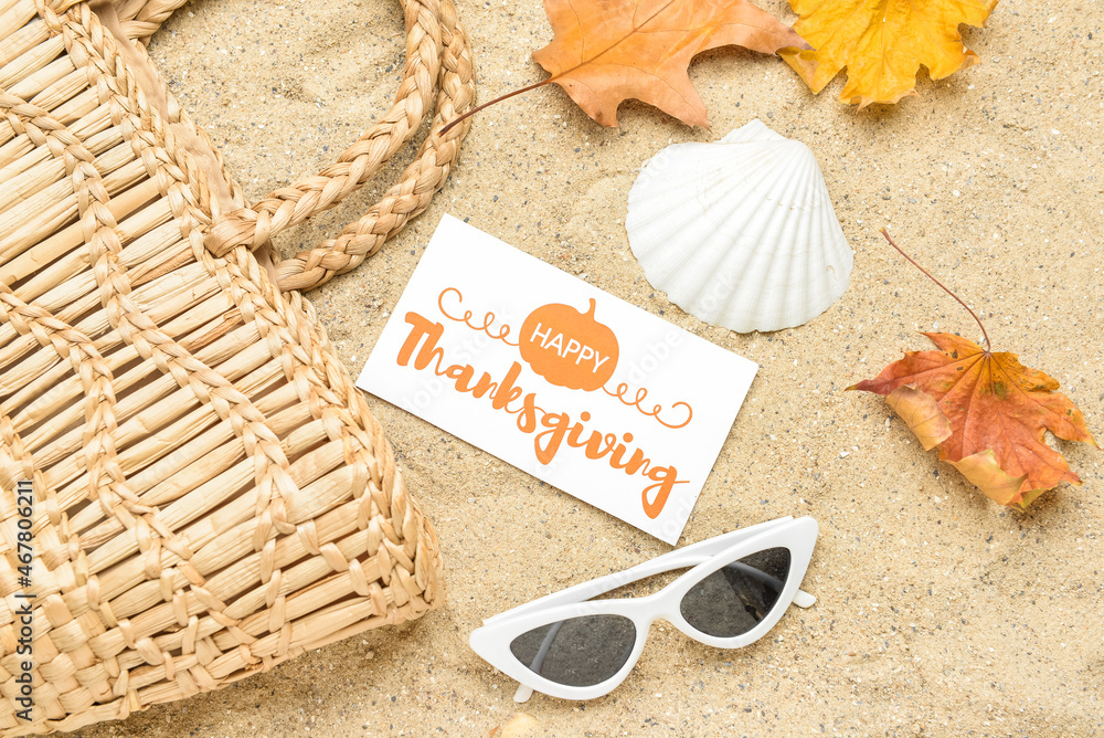 Wicker bag, dry leaves, sunglasses, seashell and paper card with text HAPPY THANKSGIVING on sand