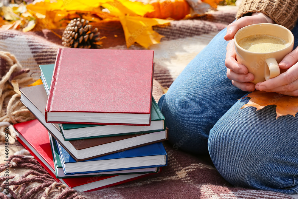 秋天公园里，一个拿着咖啡的女人坐在一摞书旁边的格子上