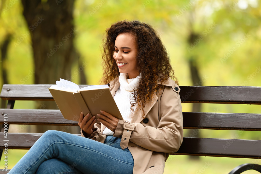 微笑的非裔美国妇女在秋季公园的长椅上看书