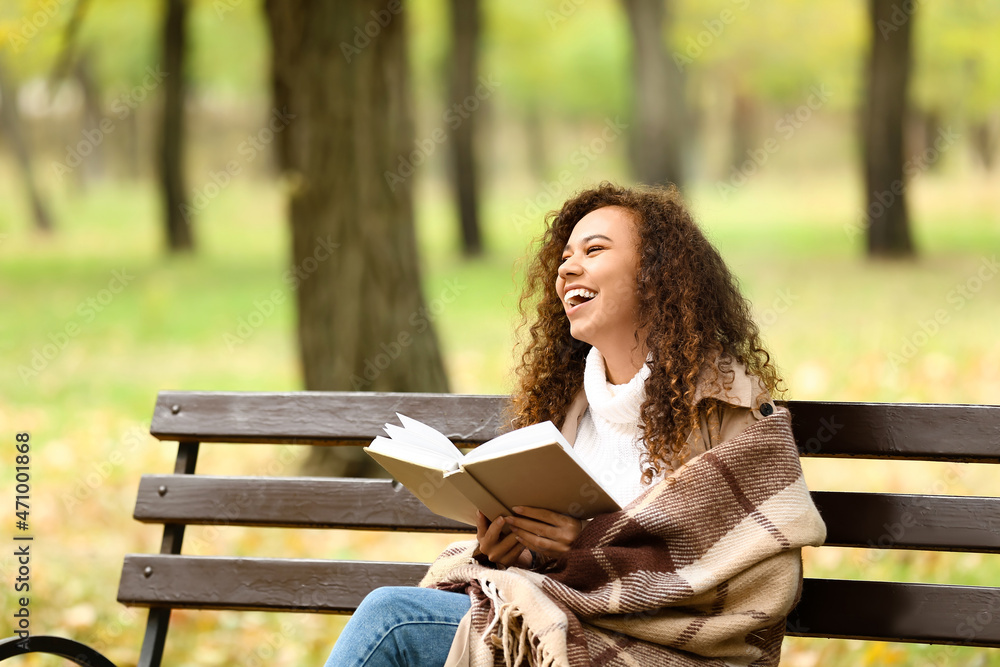 快乐的非裔美国女人在秋天公园的长椅上看书