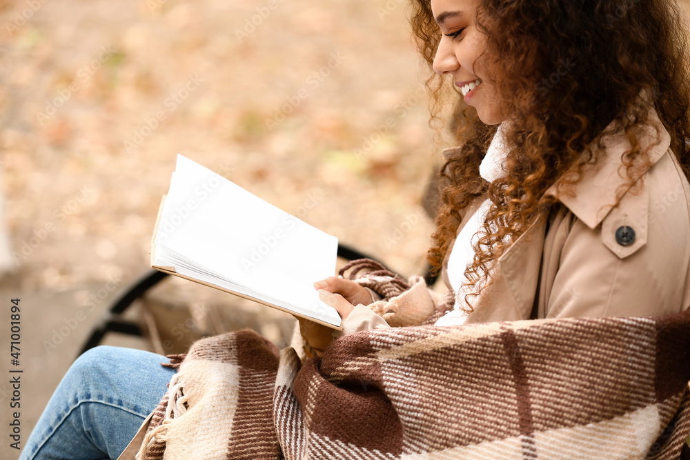 微笑的非裔美国妇女在秋天公园的长椅上穿着温暖的格子看书