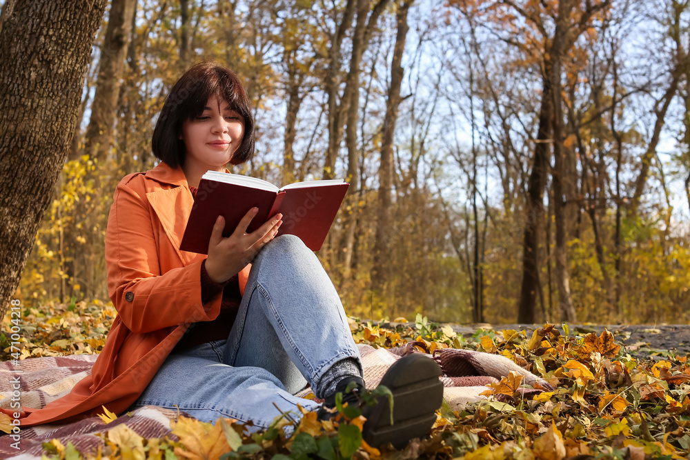 年轻女子在美丽的秋季公园里坐在格子布上看书