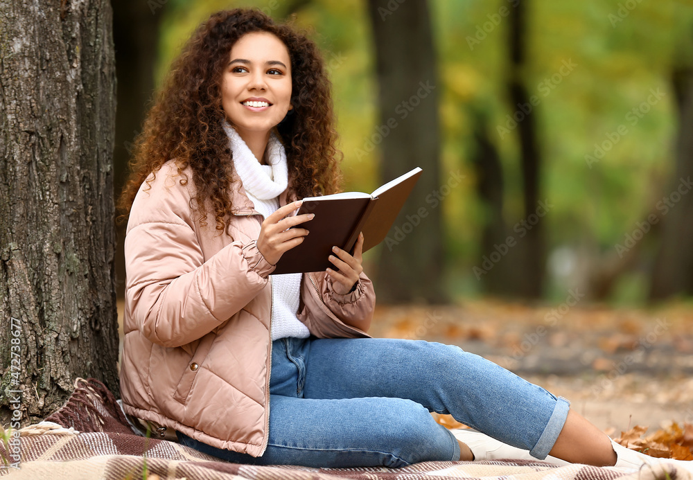 秋天公园里，一位年轻微笑的非裔美国妇女坐在格子布上看书