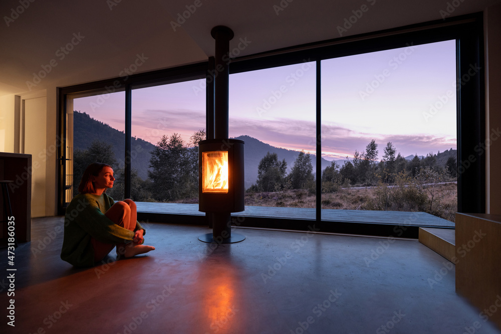 黄昏时分，女人坐在现代客厅的壁炉旁，欣赏群山美景。Conc