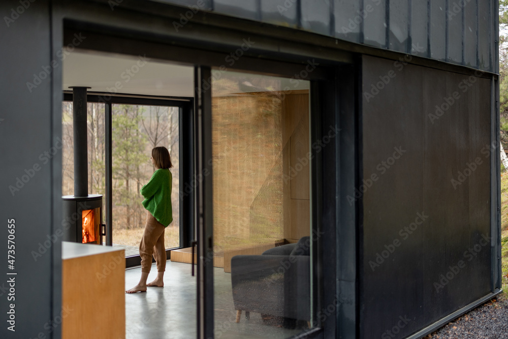 Young woman stands near fireplace at modern house on nature. View from outside through the window. C