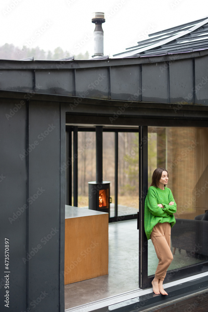 年轻的女人站在现代房子的窗户前，看着大自然。舒适和快乐的概念