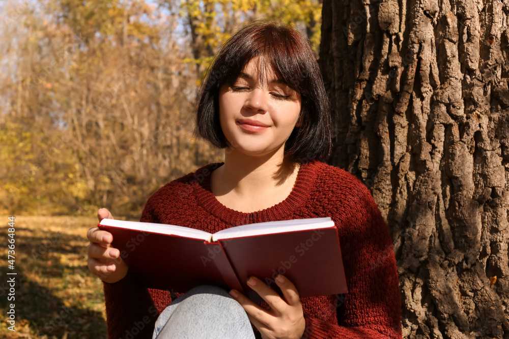 年轻女子坐在树旁，在秋季公园读有趣的书