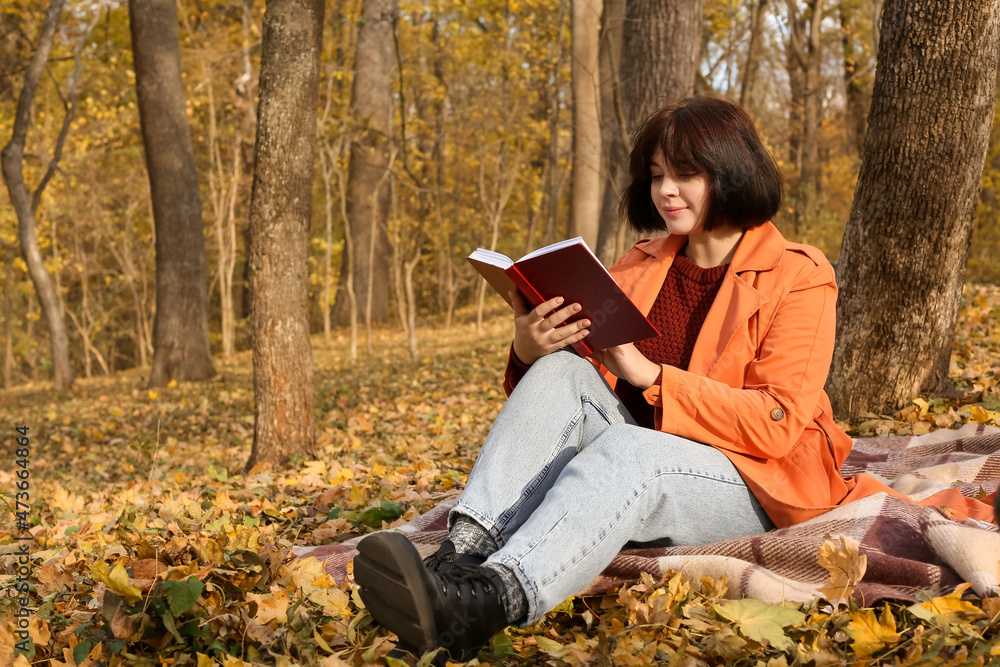 年轻女子坐在树旁的格子上，在秋季公园看书