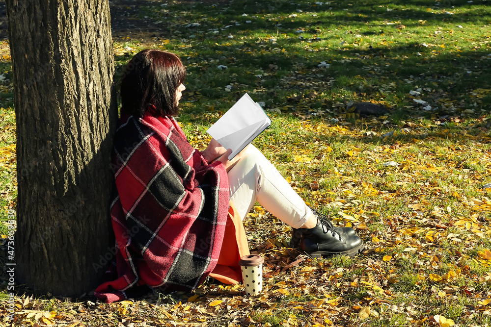 Woman sitting near tree and reading book with blank pages in autumn park