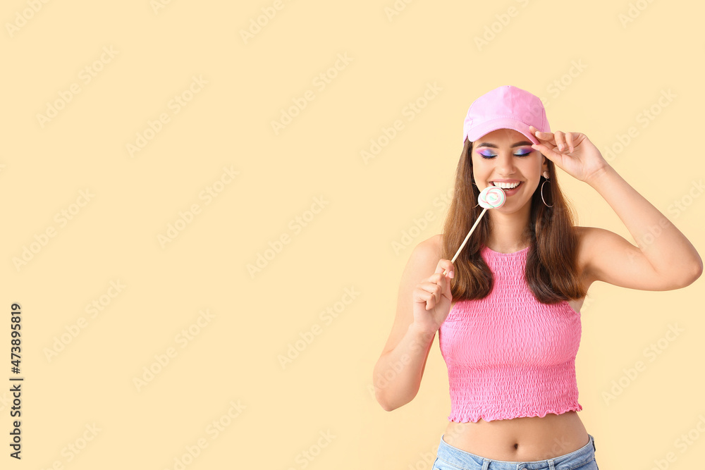 戴粉色帽子的漂亮年轻女子吃米色背景的甜棒棒糖