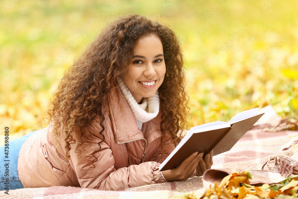 Beautiful African-American woman with book on plaid in autumn park