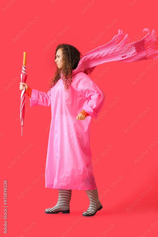 一位年轻的非裔美国女性，穿着粉色雨衣、围巾和秋葵靴，红色背景上有伞