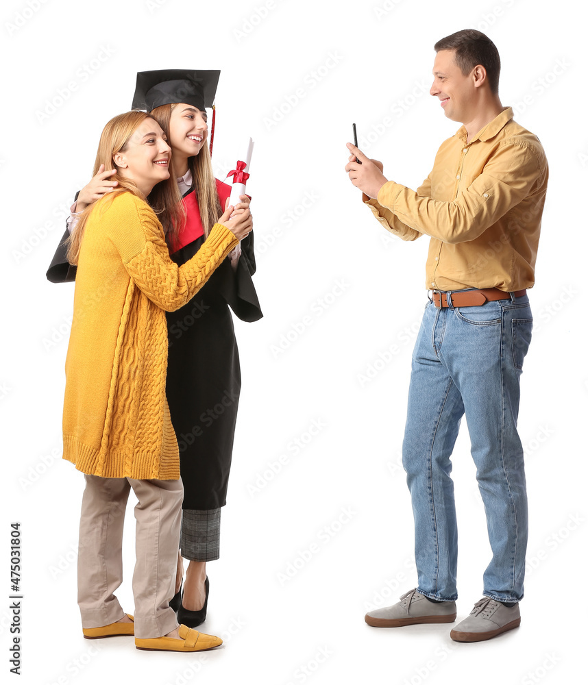 快乐的男人穿着毕业礼服和白色背景上的文凭给妻子和女儿拍照
