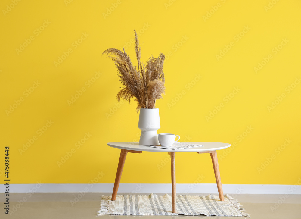 黄色墙壁附近的桌子上放着装有干芦苇、杯子和报纸的花瓶