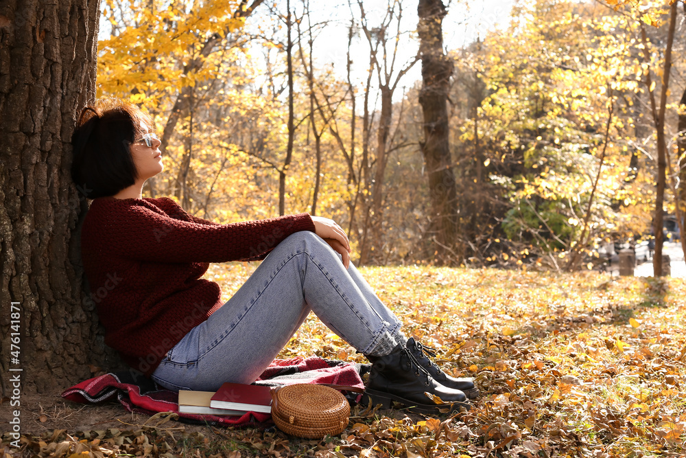年轻时尚女士坐在树旁，在公园里享受阳光明媚的秋日