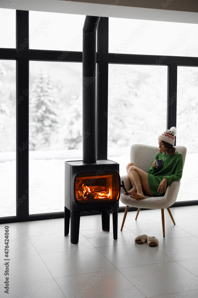 冬天，在现代自然之家的壁炉旁，一个女人拿着杯子坐在椅子上。Conce