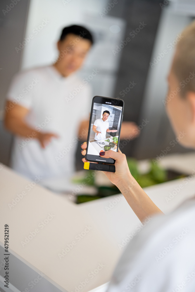 女人在智能手机上拍摄亚洲男人在厨房烹饪素食沙拉的视频。视频的概念