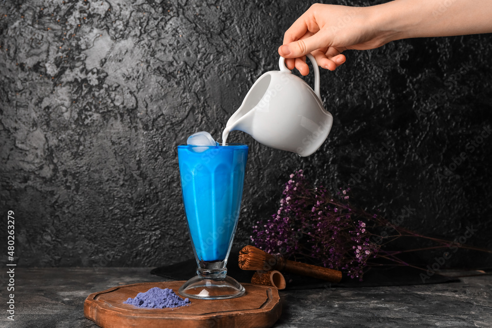 一名女子将牛奶从罐子里倒进一杯深色背景的蓝色抹茶拿铁
