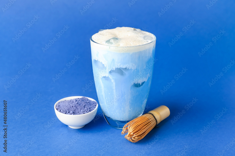Glass of cold matcha latte, powder and chasen on blue background