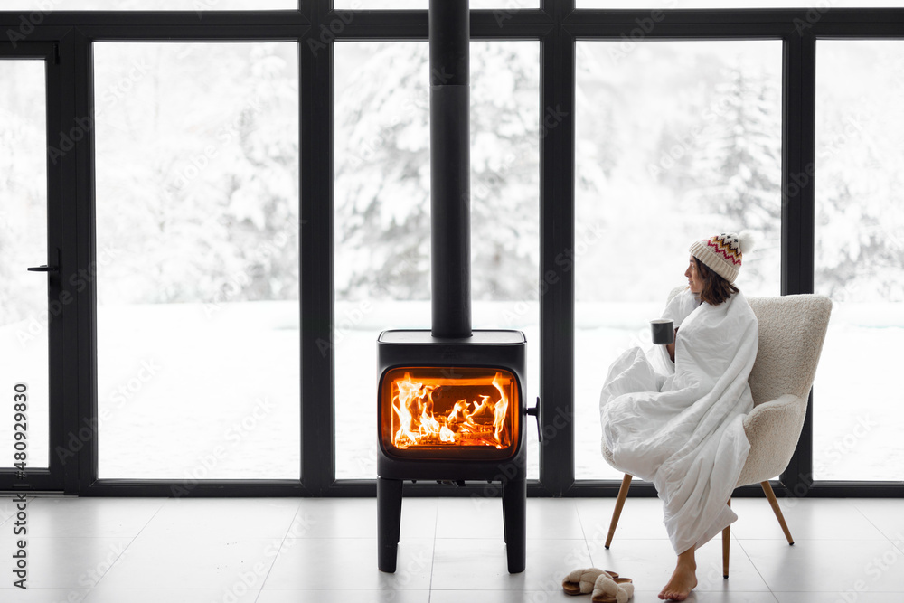 Woman covered with blanket enjoys winter time at home with burning fireplace, looking outside the wi