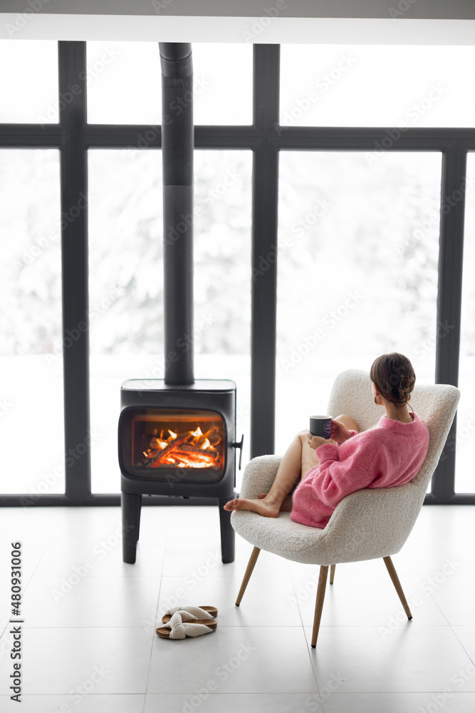 冬天，在现代自然之家的壁炉旁，一个女人拿着杯子坐在椅子上。Conce