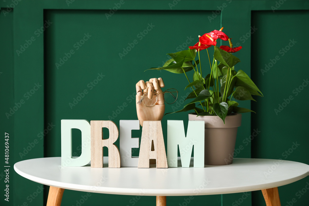 Table with Anthurium flower, wooden hand holding eyeglasses and word DREAM on color background