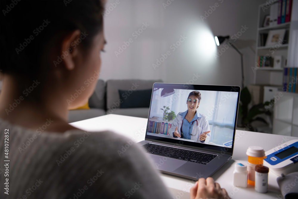 年轻的亚洲女性坐在家里的沙发上，使用无线互联网与医生进行视频通话
