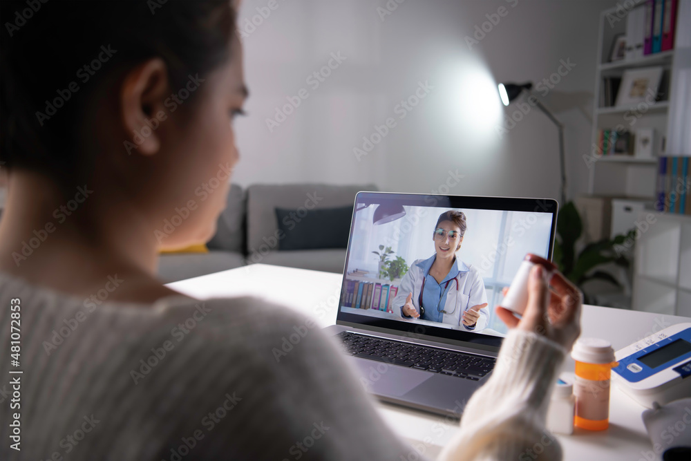 年轻的亚洲女性坐在家里的沙发上，使用无线互联网与医生进行视频通话