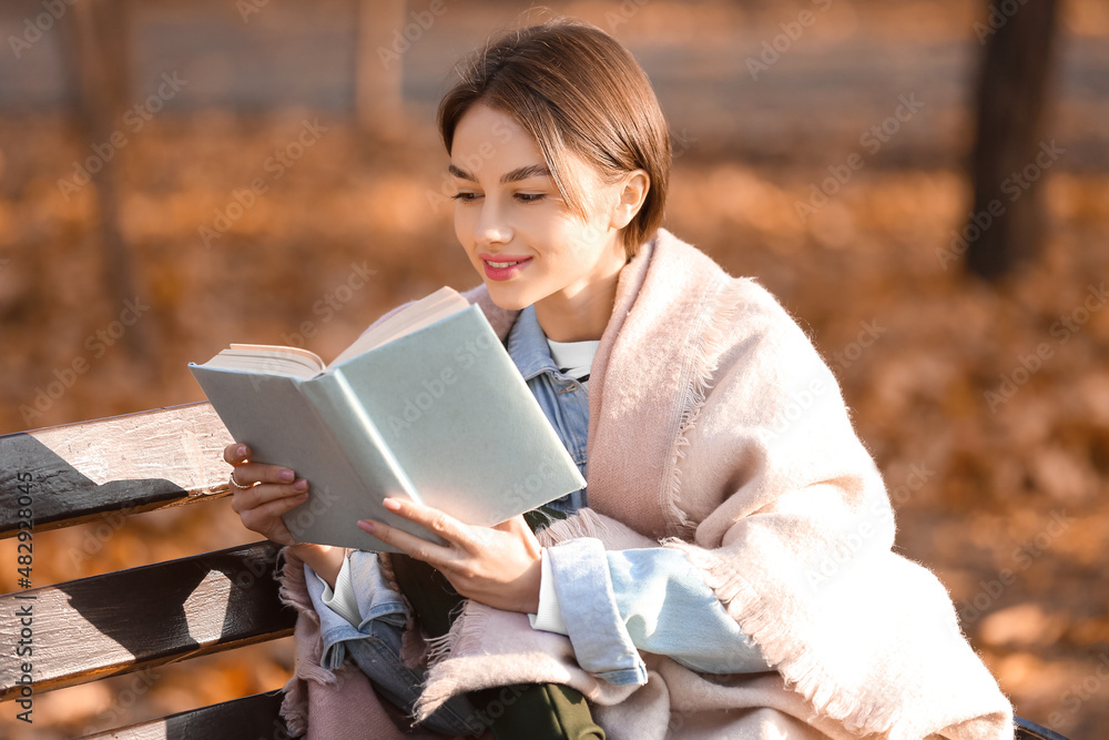 秋天公园的木长椅上有一位年轻的女士，她拿着格子看书