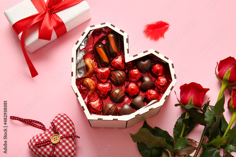 Heart shaped box with tasty chocolate candies, roses and gift on pink background