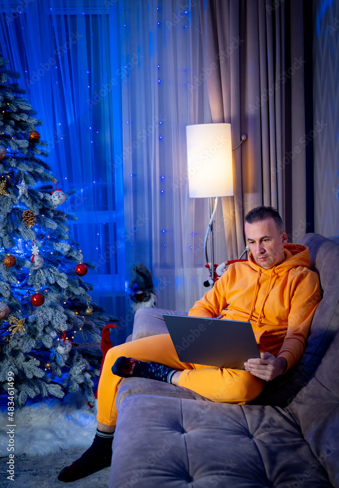 Man wearing pajama sits on a sofa behind a laptop on the background of New Year room interior. Man w