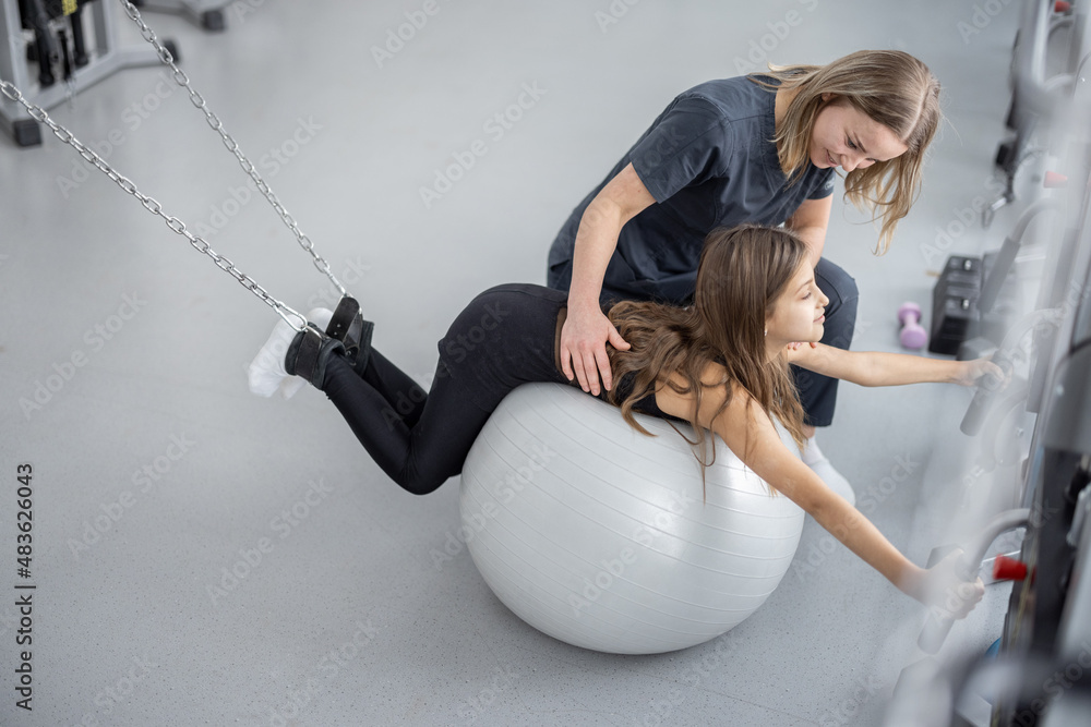 Rehabilitation specialist helping little girl to do exercises with fitness ball and decompression si