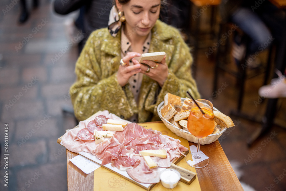 坐在餐厅里，一位女士正在拍摄一个装有意大利肉类开胃菜和雪碧Aperol饮料的盘子