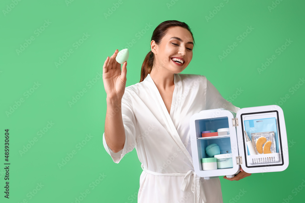 Happy young woman with open cosmetic refrigerator and sponge on green background