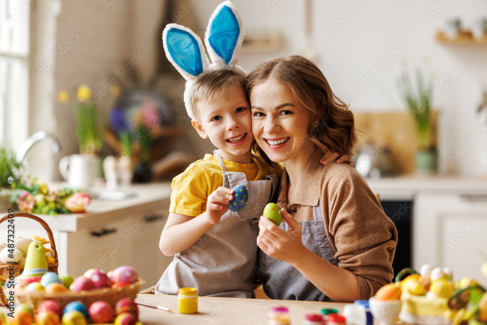 Loving young mother teaching happy little kid son to decorate Easter eggs while sitting in kitchen
