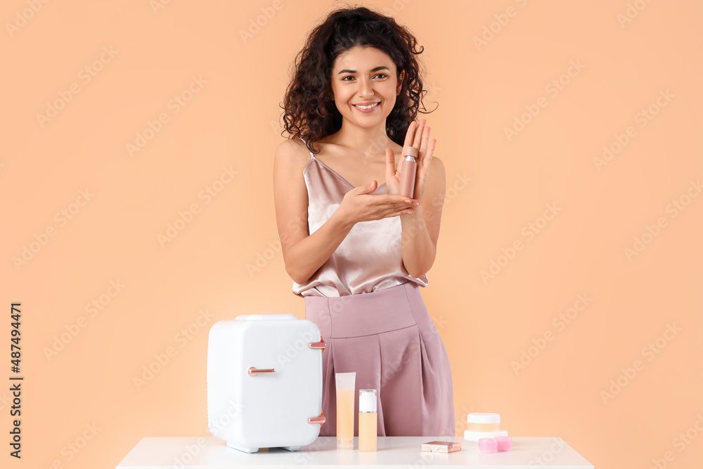 Beautiful smiling woman holding bottle of perfume near table with refrigerator and cosmetic products