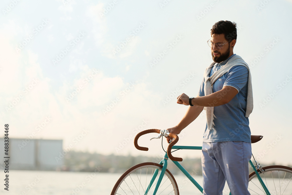 年轻的胡子男子骑着自行车在路堤上看手表