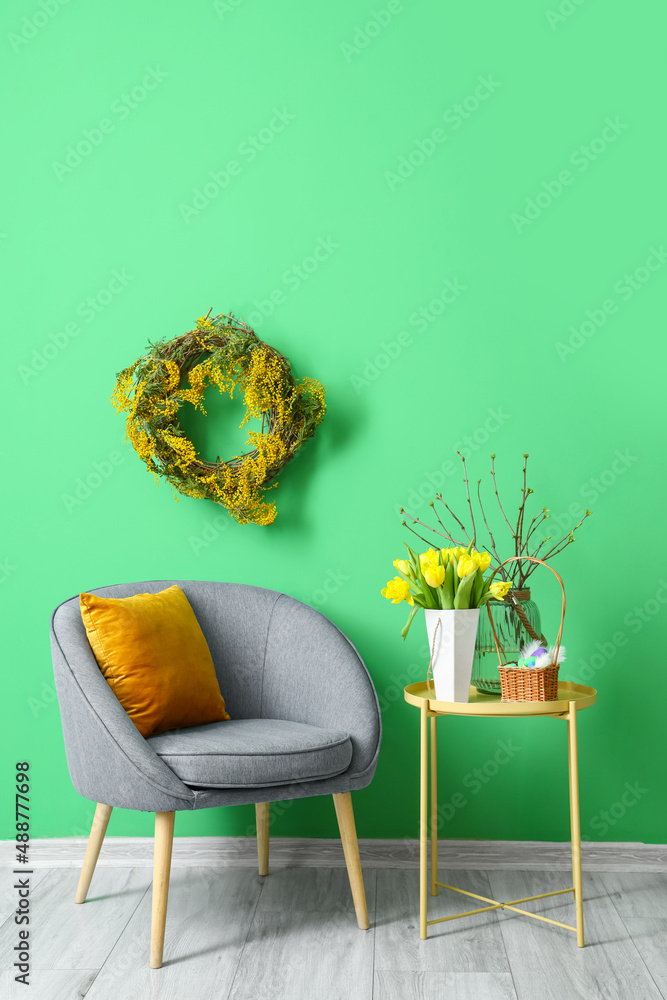Comfortable armchair, table with tulips and basket of Easter eggs near color wall