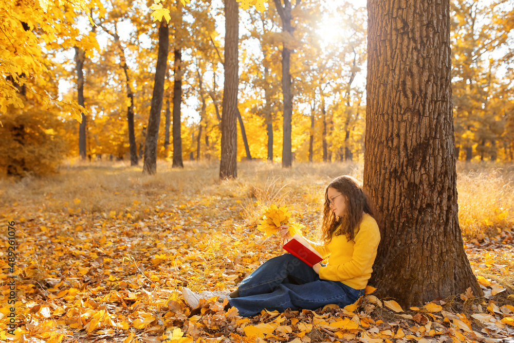 Woman holding bouquet of autumn leaves and reading book near tree in park