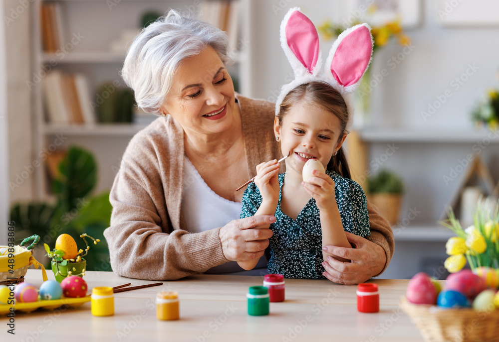 慈爱的祖母教快乐的小女孩坐在厨房里装饰复活节彩蛋
