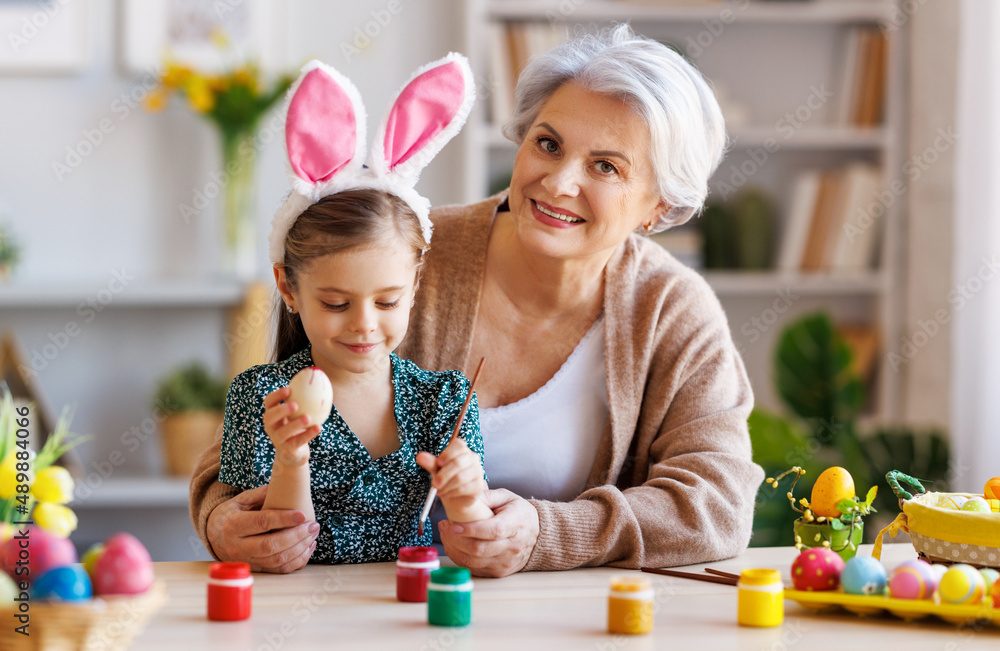 Loving grandmother teaching happy little kid girl to decorate Easter eggs while sitting in kitchen